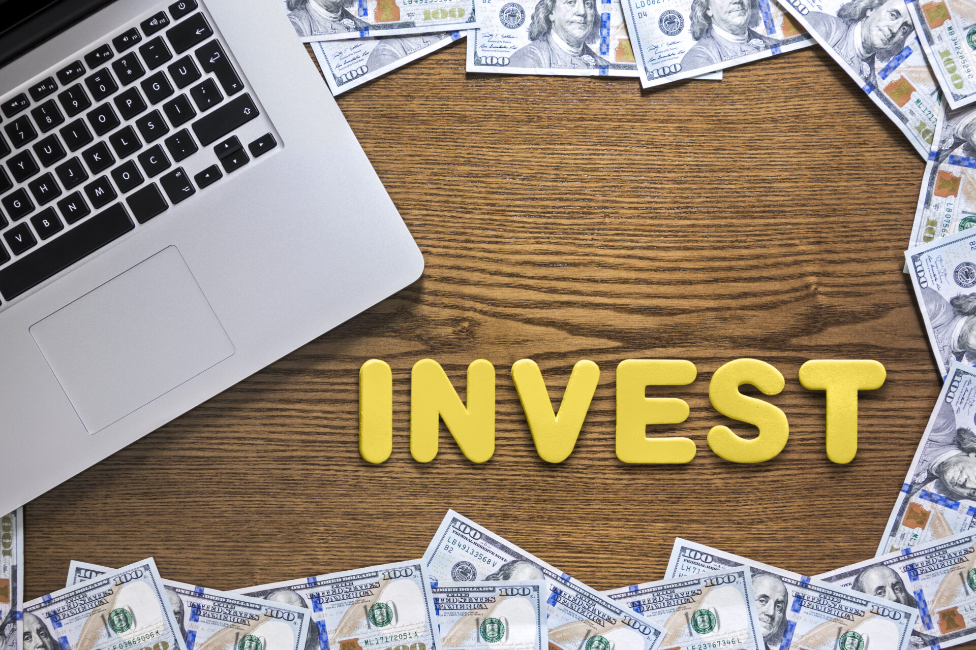 Looking down on wooden desk with laptop and 100 dollar bills with “Invest” text